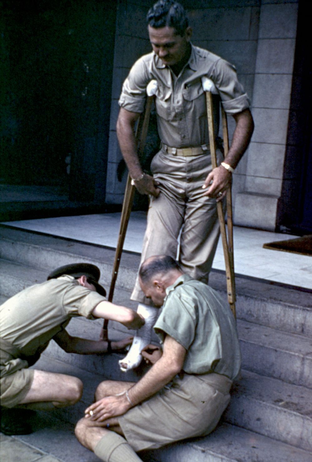 Colour photograph of 2 servicemen sign the leg cast of War correspondent Earl Crotchett on crutches on the steps of the Queens Hotel in Townsville 1942