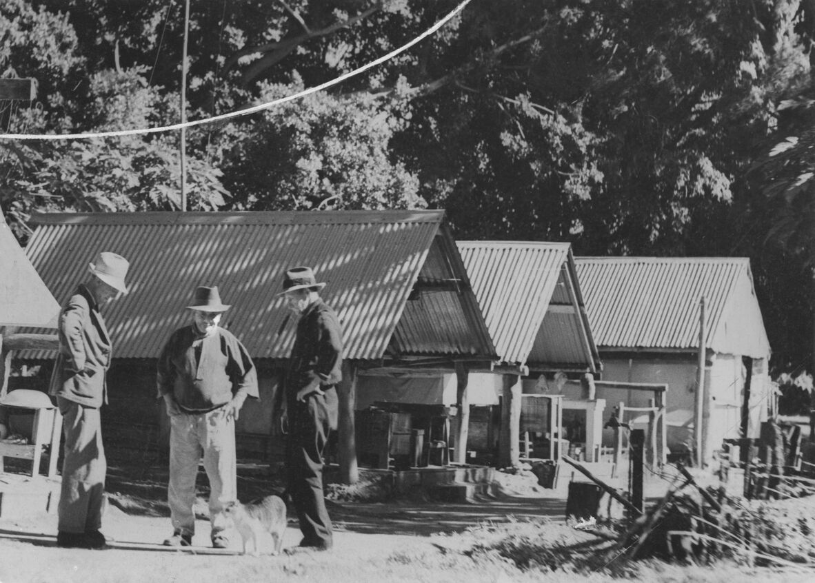 Black and white photo of three men talking outside a row of huts with corrugated iron roofs