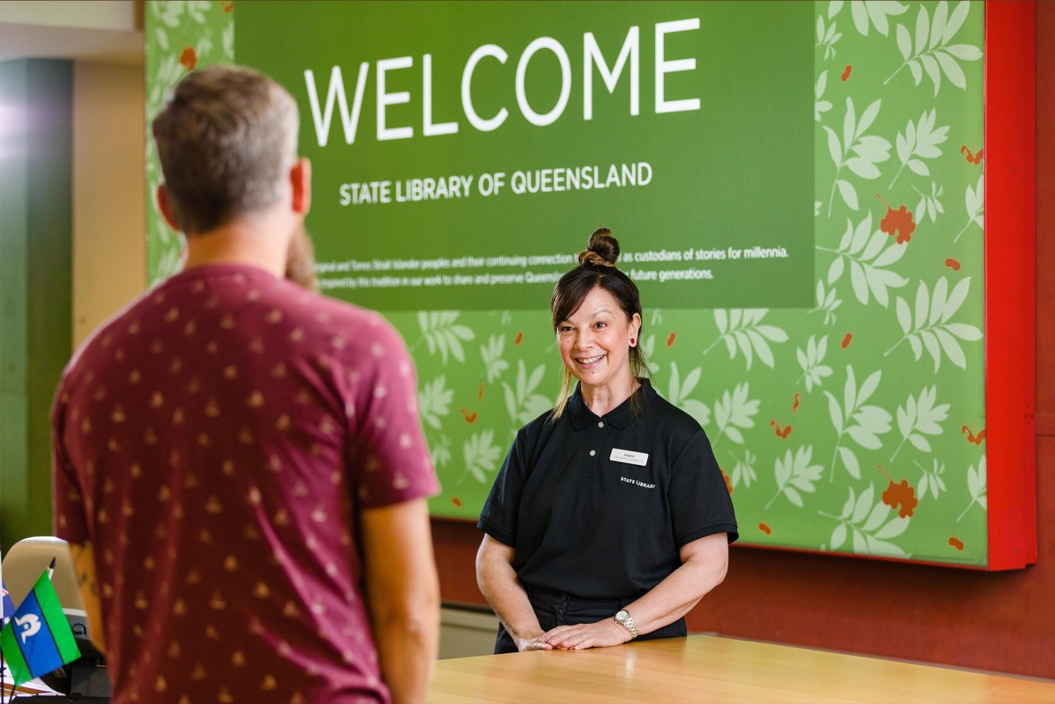 Client facing a smiling State Library staff member at the reception desk The wall behind the staff member says Welcome