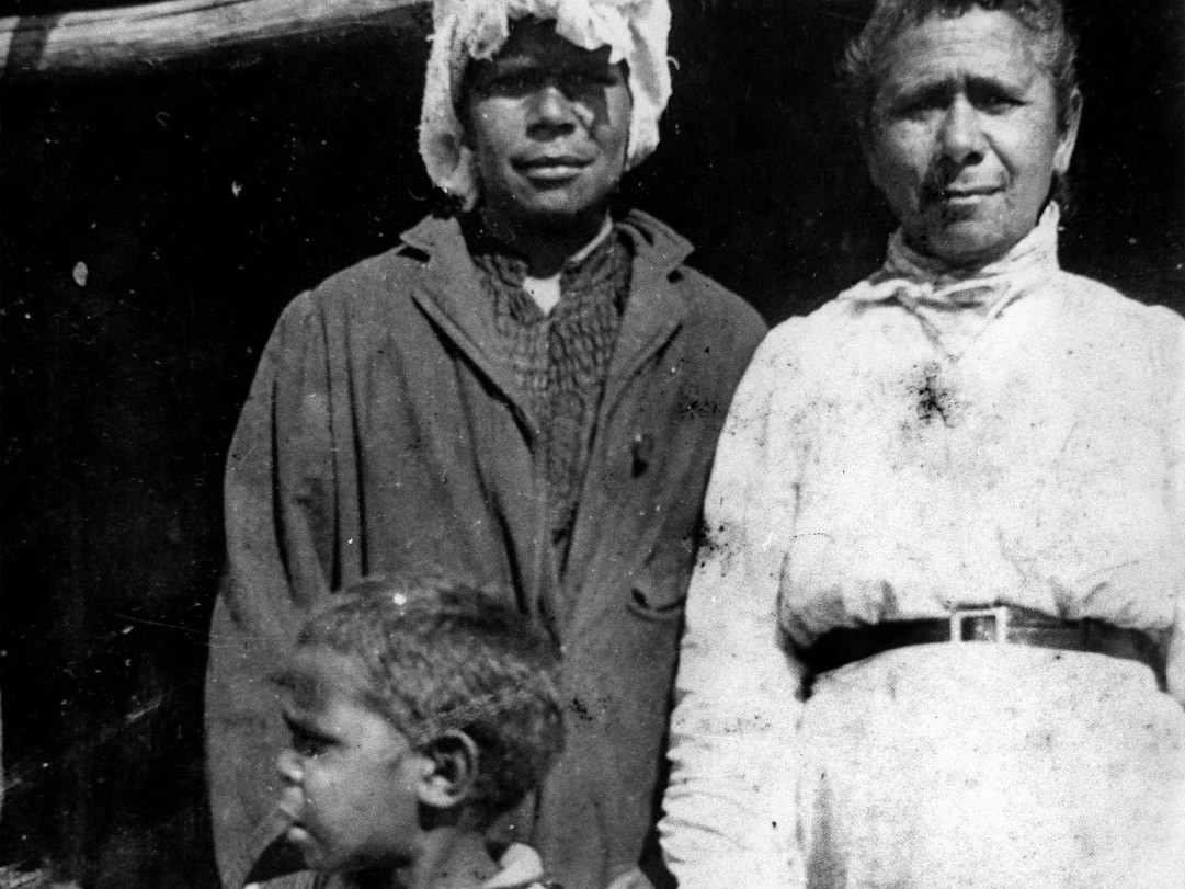 Black and white image of two women standing in front of a building, with a child standing in front of them, ca. 1905