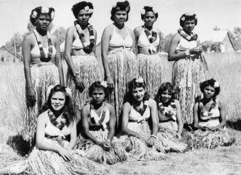 Group of First Nations women in bikini tops and grass skirts in a field 5 standing and 5 sitting