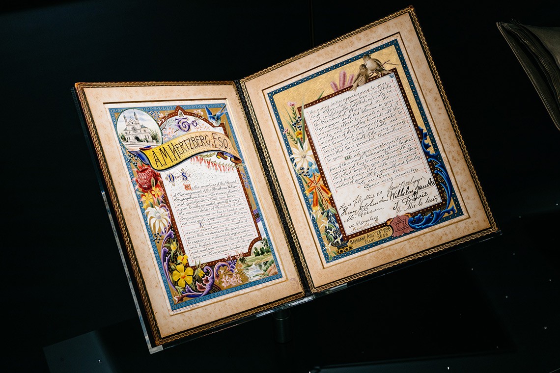  An illuminated address presented to Adolphus Hertzberg President of the Brisbane Hebrew Congregation in 1896  The illuminated address is contained in an olive coloured leatherbound gold embossed album with silver insignia  The inner pages feature an illustration of the Brisbane Synagogue and borders containing birds and flowers decorated with gold and silver