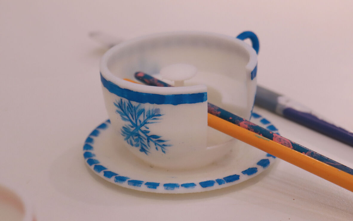 Image of of tiny, white 3D printed tea cup and saucer and paint brushes. The cup and saucer have hand painted traditional blue decorations.