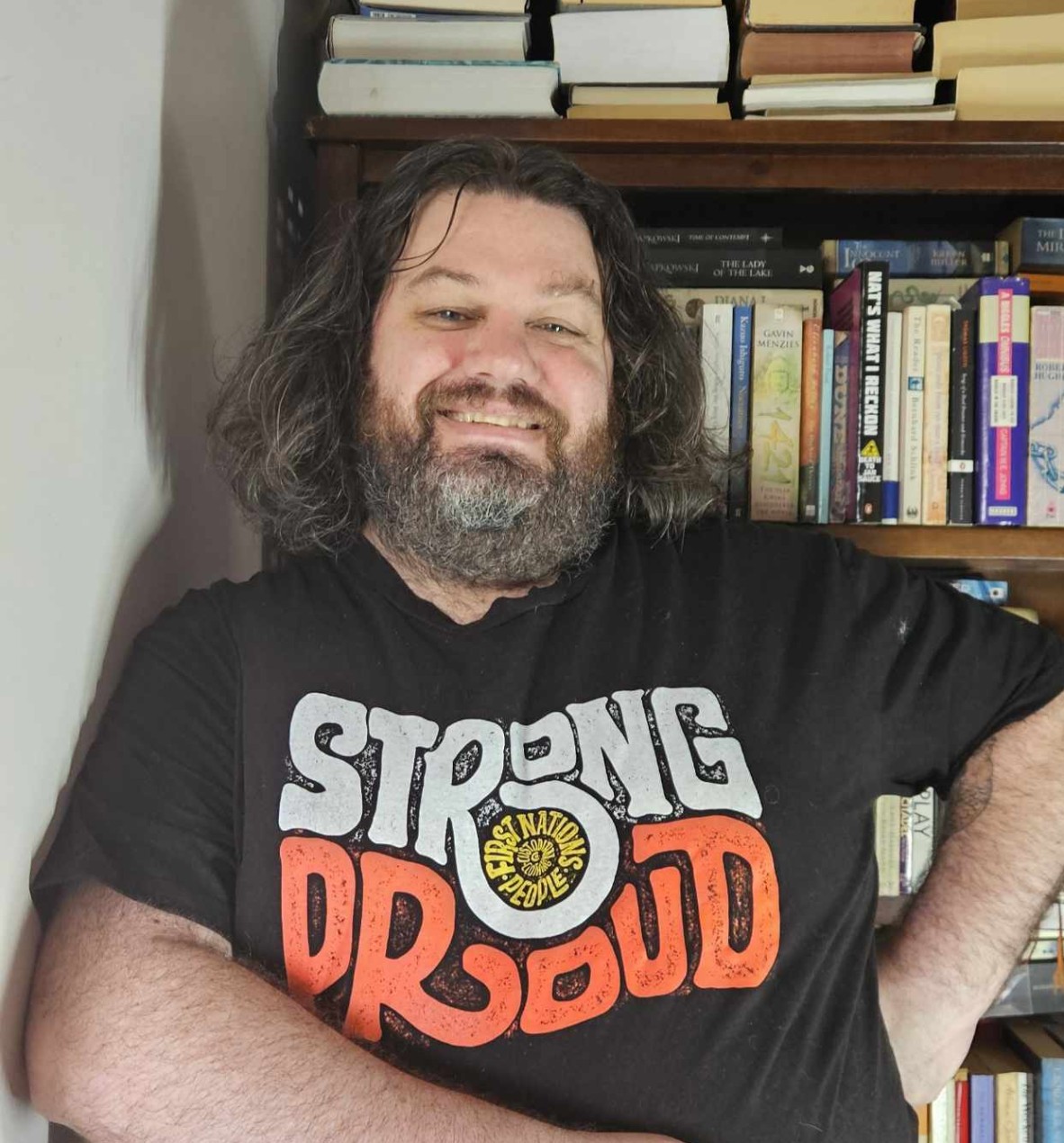 A smiling man in a black t shgirt standing in front of a tall bookcase. The shirt has writing which reads strong and proud.