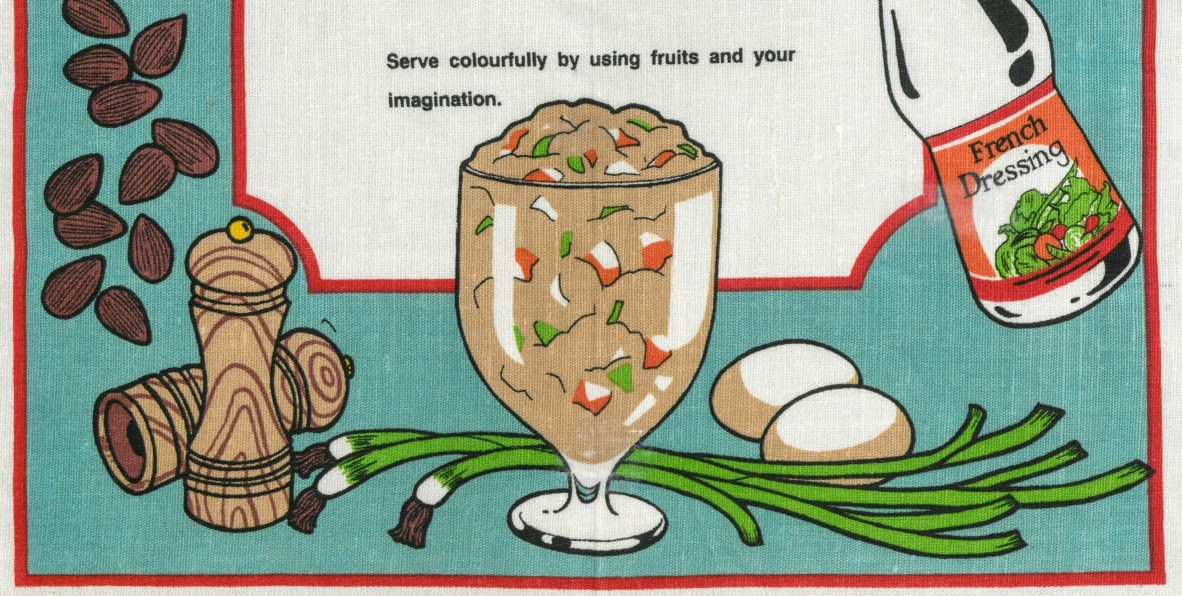 Detail of crayfish and chicken salad recipe on teatowel. A tall glass vessel shaped like a sundae or milkshake glass is filled with lumpy brown salad. around the edges, almonds, sald and pepper shakers, a french dressing bottle, french onions, and two eggs are scattered. A red decorative border frames the blue background.
