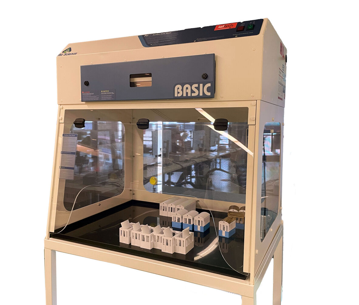 A laboratory fume hood labeled 'Basic' on display in a workspace. Inside the fume hood are small architectural models of buildings with white walls and blue bases. The background shows a modern workspace with desks and computers.
