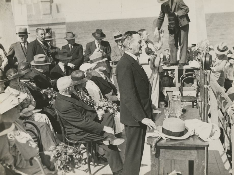 The Governor Sir Leslie Orme Wilson stands to make a speech at opening of the Hornibrook Highway also broadcast on radio Manuel Hornibrook his wife Daphne and other dignitaries sit behind him with journalists in the background