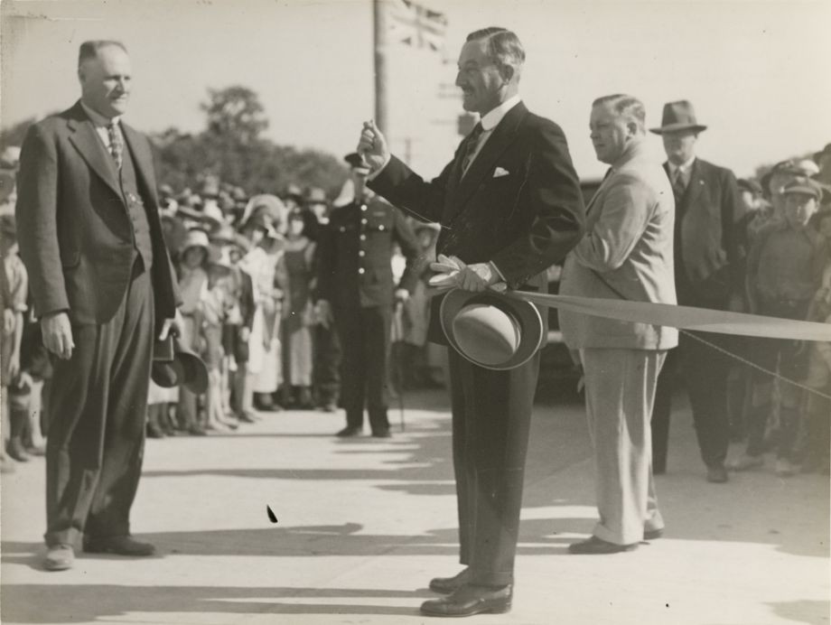 This image looks like the moment when the ribbon has been cut to open the Hornibrook Highway, by the Governor, Sir Leslie Orme Wilson. Standing opposite him is Manuel Hornibrook in a proud moment. Manuel gave the Governor a gold boomerang to mark the occasion, which was used to cut the ribbon.