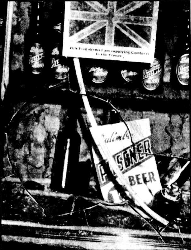 High-contrast black-and-white photograph of a damaged shopfront window with a large crack spiderwebbing across the glass. Behind the cracked pane, a display features several bottles of Pilsner beer lined up on a shelf. A white card depicting the Union Jack flag stands prominently in the center, with the text underneath reading: "This Flag shows I am supplying Comforts to the Troops." Below, a fallen, rectangular sign reads "Pilsener BEER" in bold lettering next to a dark glass bottle on a lower shelf.