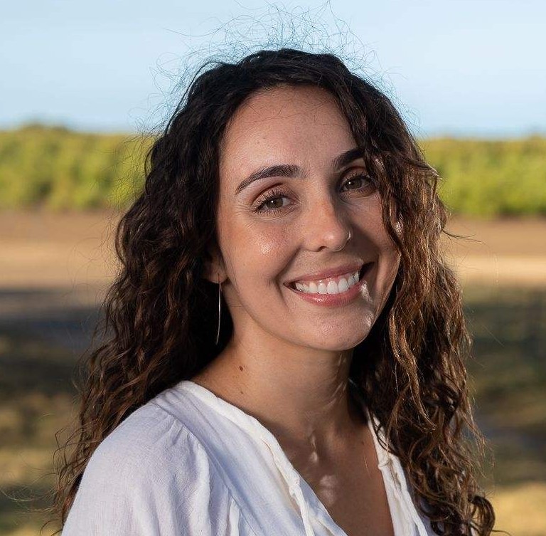 A smiling woman in a white shirt standing in front of a landscape with a bright blue sky.