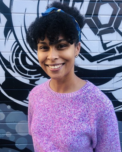 Jasmin McGaughey is wearing a pink jumper and standing against a black and white brick wall. She is smiling.