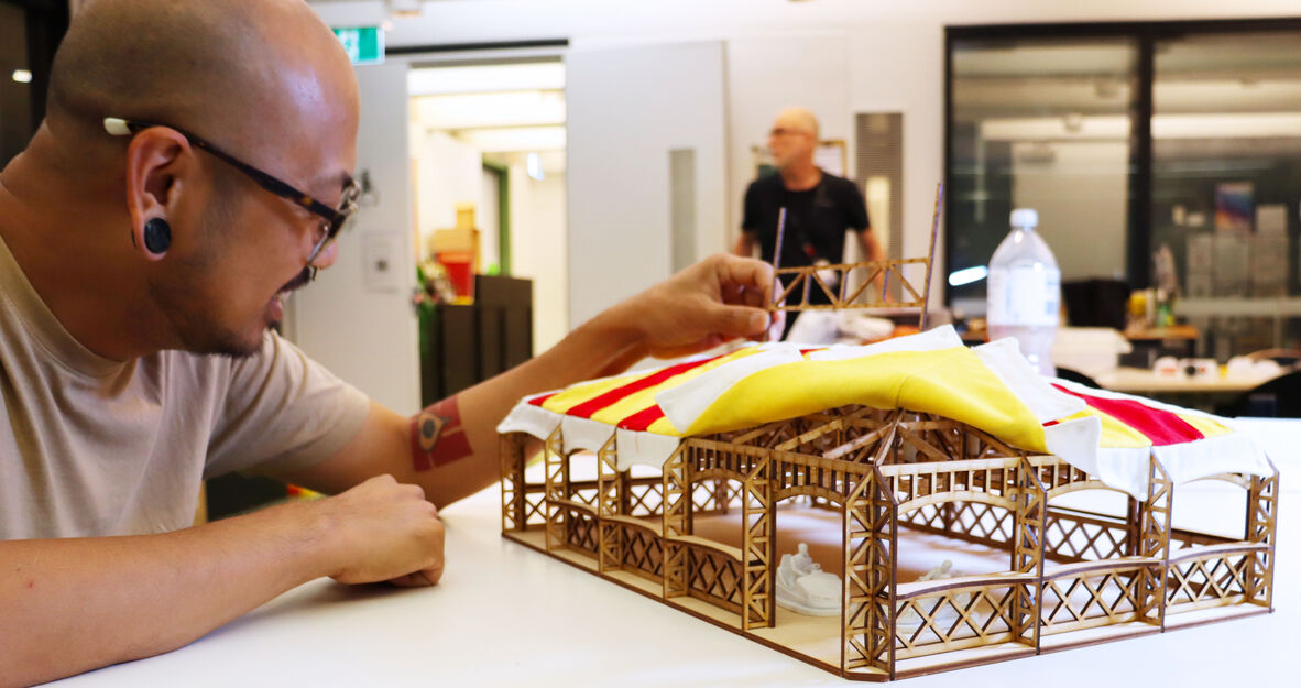 A man with glasses is reaching out and touching a laser cut wooden frame that is covered in a red and yellow fabric. The man is smiling.