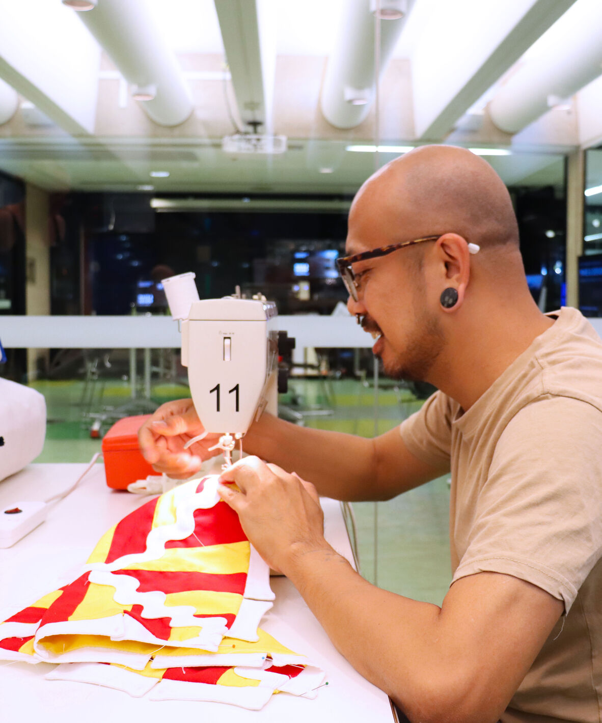 A man with glasses and piercings is sitting at a white table and sewing a red and yellow tent roof cover on a sewing machine, he is smiling while working on this.