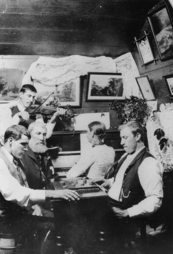 Black and white image of 3 men sitting around a checkers board, 1 man standing playing violin and woman playing upright piano in backgound, ca.1907.