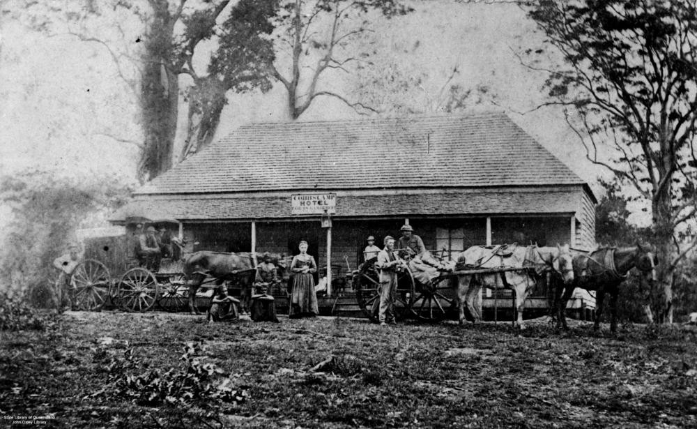 black and white image of people and horse drawn carts stand outside a hotel in woombye