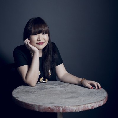 Portrait of Julie Koh sitting at a table. She wears a black shirt and necklace, and rests her chin in her hand.
