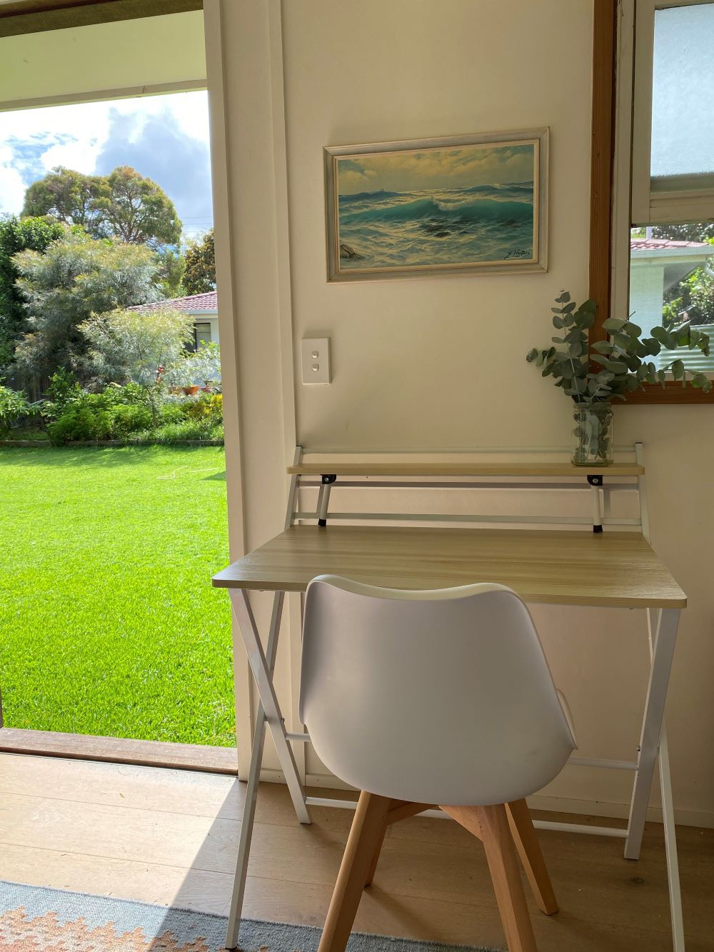 Kays workspace at home - a white chair and desk with a plant sit beneath a painting Theres a view of a green lawn