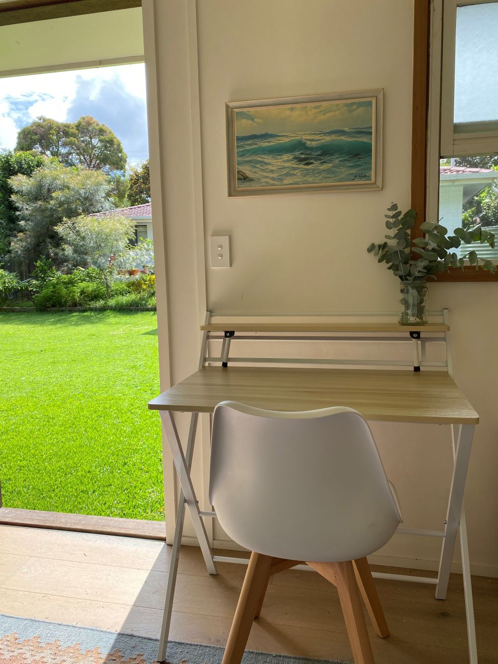 Kay's workspace at home - a white chair and desk with a plant sit beneath a painting. There's a view of a green lawn.