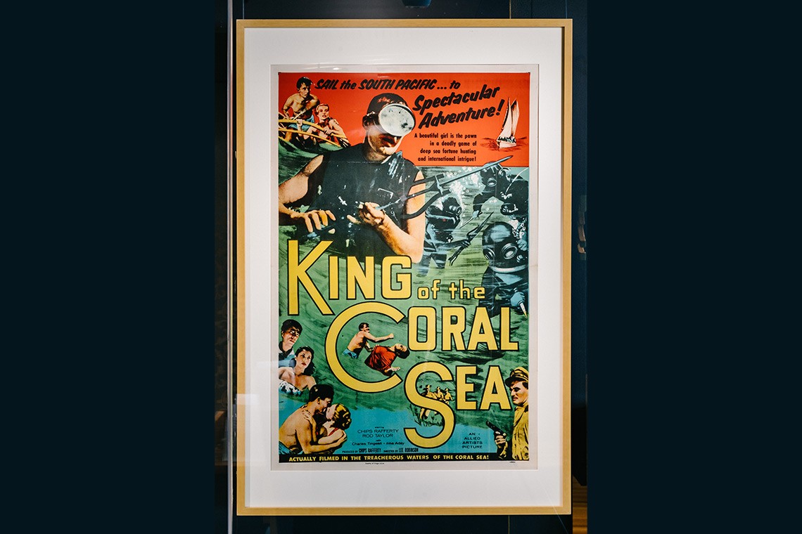 Colour lithograph poster promoting the 1954 Australian film King of the Coral Sea which was shot on location on Thursday island and in the waters off Green Island near Cairns  The coloured poster features illustrations of a sailing boat deep sea divers wearing helmets a man in a scuba mask holding a spear gun and a man and woman in swimsuits kissing passionately  