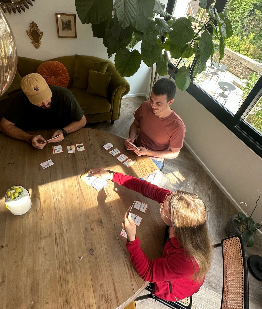 Three people sitting around a light coloured round table playing the cooked game, three people are playing and there are a bunch of cards in the table with each person holding a bunch of cards in their hands. 