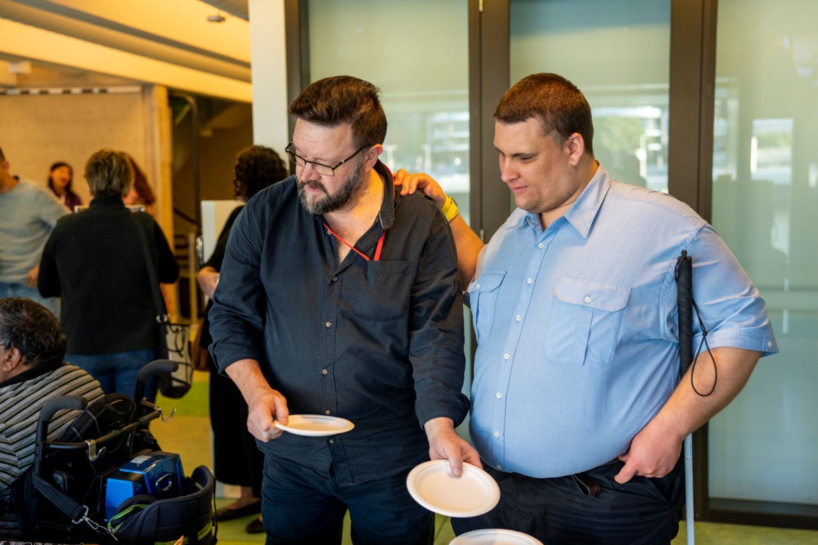 Brendon and Mark enjoy afternoon tea. Brendon has a blue shirt and holds his cane by his side. Mark has a dark shirt and a beard. He helps Brendon to get a brownie to eat.