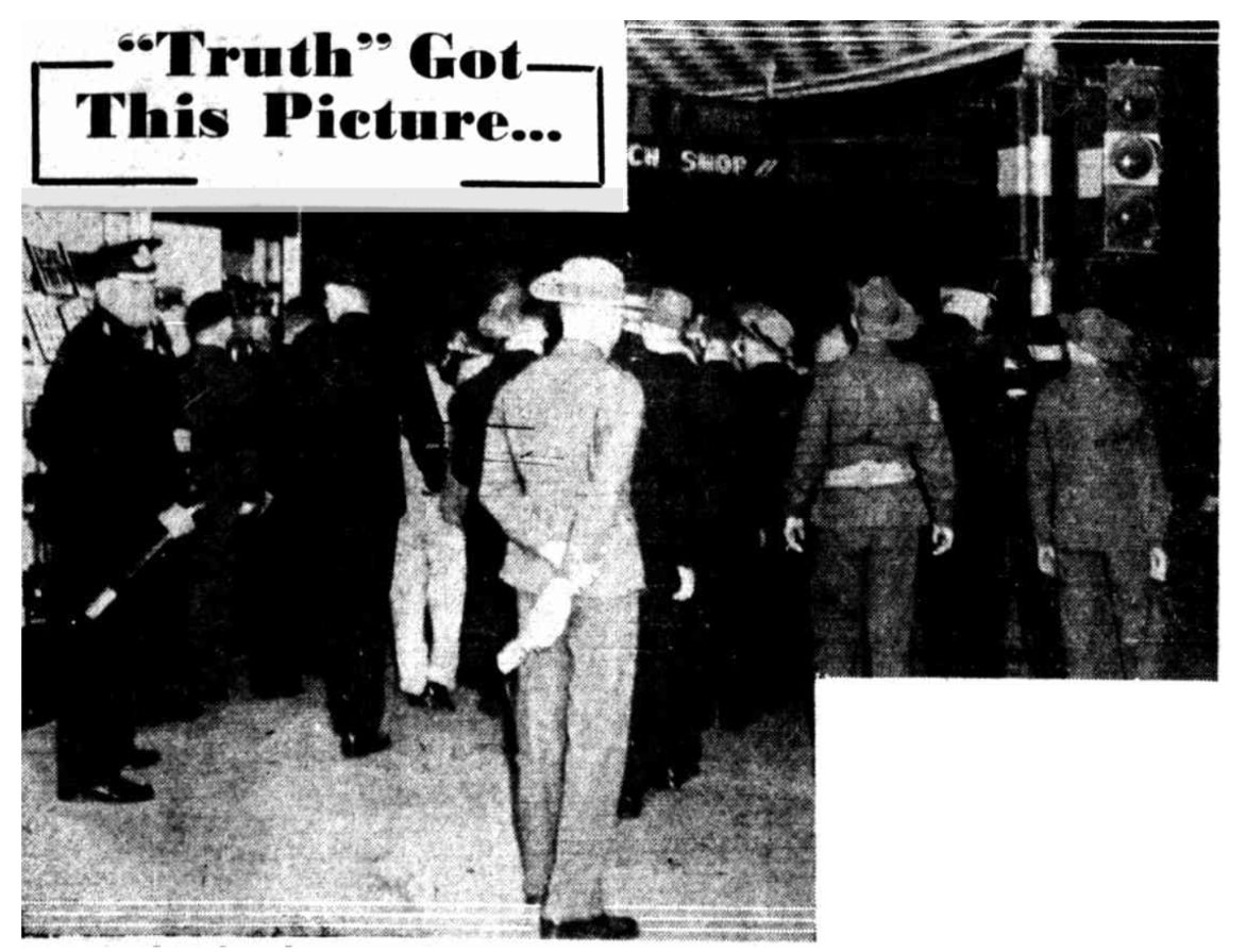 An archival, high-contrast, black-and-white photograph shows a crowd of military and police personnel gathered on a street at night. In the foreground on the left, a police officer in a dark uniform and peaked cap stands facing the crowd, holding a baton in his hand. Near the center, a man in a light-colored military uniform with a wide-brimmed slouch hat stands with his back to the camera, his hands clasped behind him. To the right, other men in military uniforms and slouch hats are seen from behind, fac