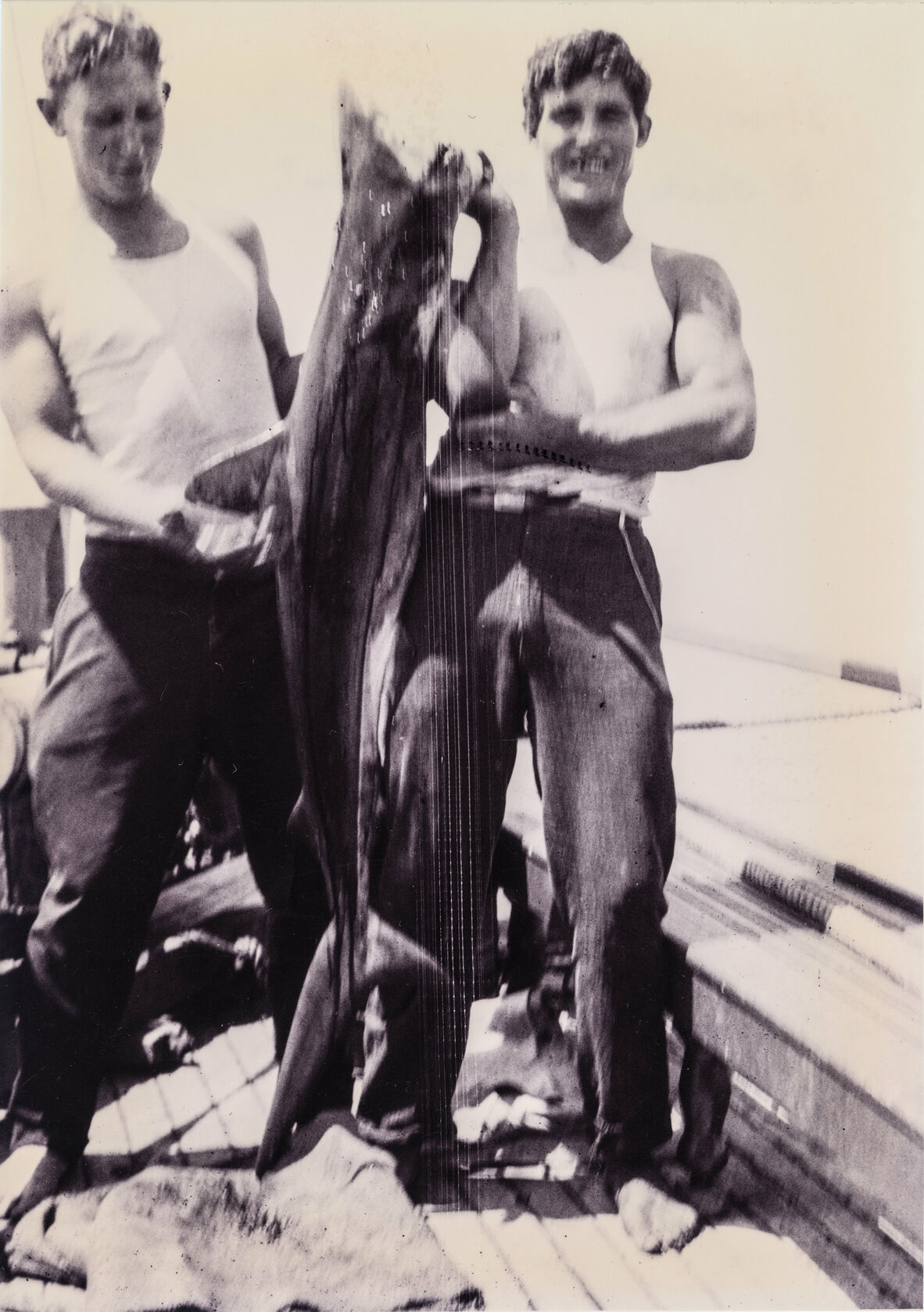 A blurry black and white photo of two men in white singlets holding up a shark they've just caught