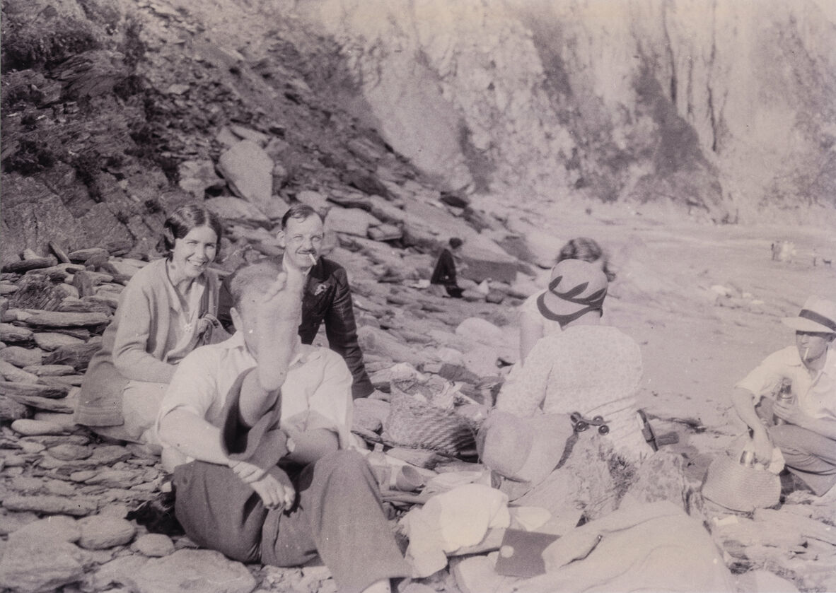 A black and white photo of six people on a rocky beach; some are smoking, eating, smiling at the camera
