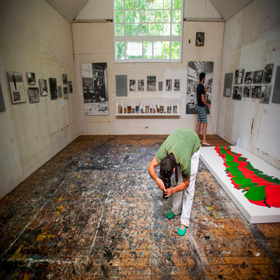 People stand around an art studio with high ceilings and pictures on the wall. A person takes a photo of the paint-marked floor.