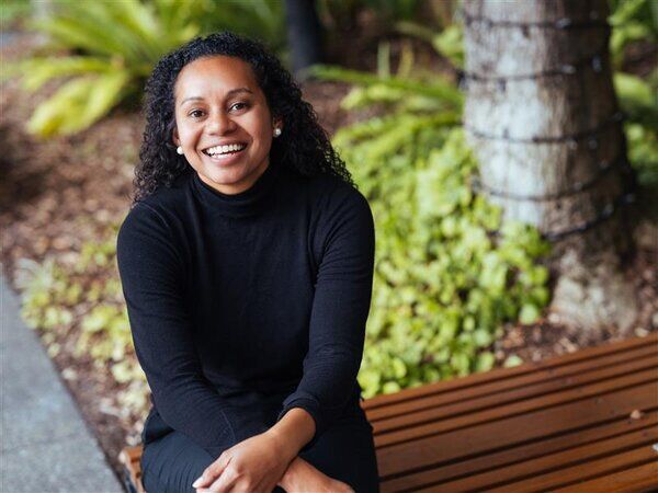A smiling woman with brown skin wearing a black top sitting on a bench with a garden background.