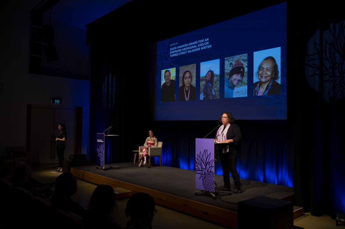 Two women are on stage during a ceremony. Five headshots are on a blue screen behind them