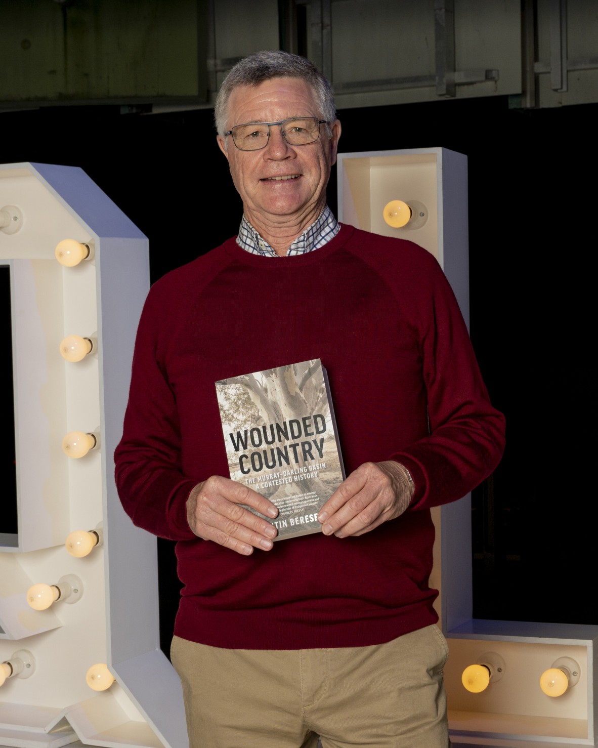 Quentin Beresford wears a red jumper and glasses. He holds up a copy of his book, Wounded Country, with lights behind him