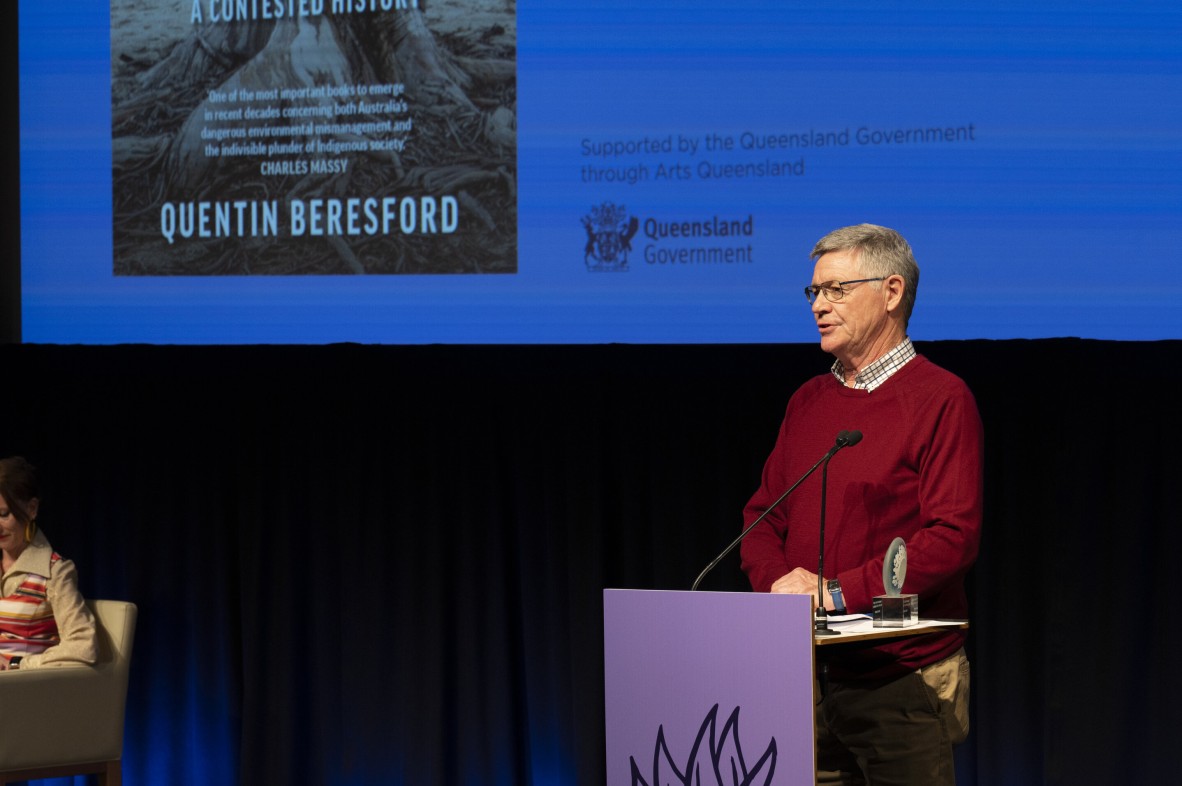 Quentin Beresford is onstage at the Queensland Literary Awards speaking behind a lectern. A screen is behind him with his book on it