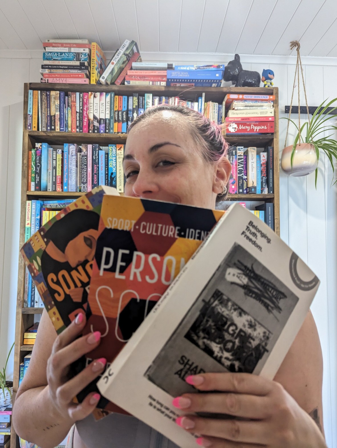 Melanie Saward smiles in front of a bookshelf. She is holding three books towards the camera.