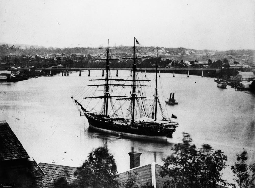 Black and white photo of 3 masted vessel Indus on Brisbane river with rooftops in foreground and bridge in background ca1874