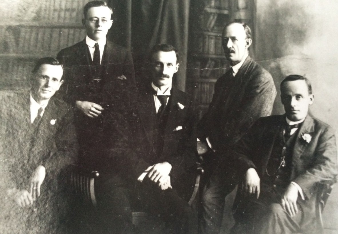 Public Library of Queensland staff, ca. 1925. Five men sitting together wearing dark suits, and two with moustaches.