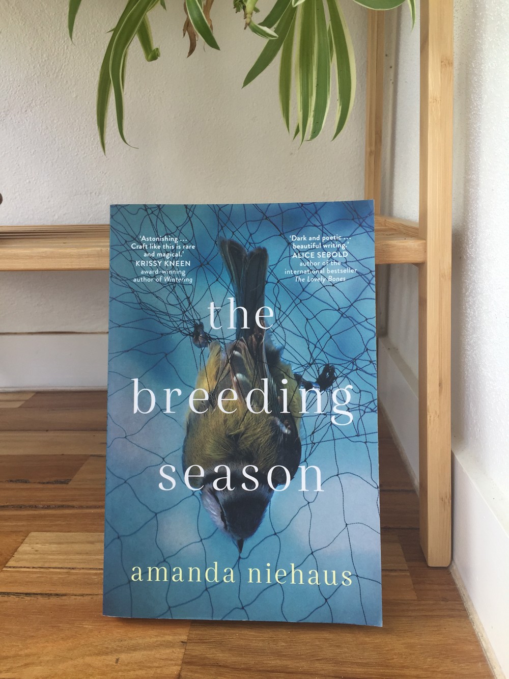 The cover of The Breeding Season by Amanda Niehaus; the cover is blue and shoes a green and grey bird caught in netting