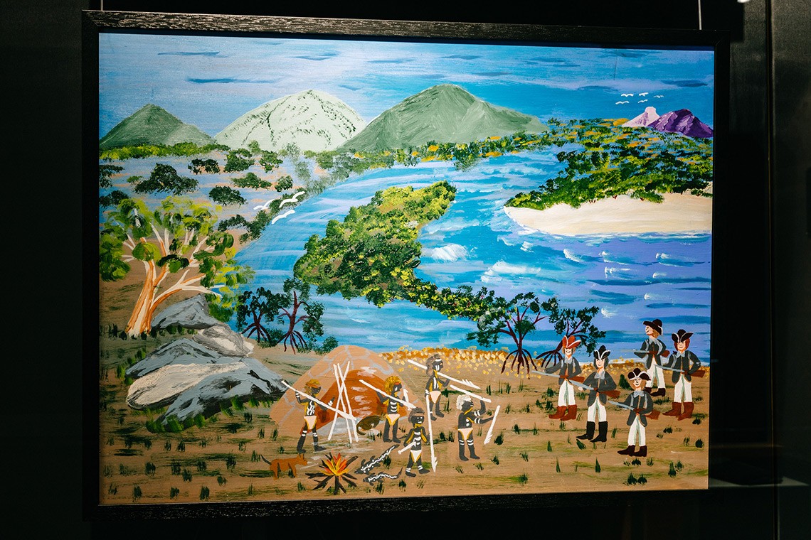   The painting Defending Our Camp depicts the First Nations oral history of Captain Cooks landing at Endeavour River  The acrylic on plywood painting depicts the landscape of the Endeavour River with the green mountains in the background and the blue river winding through the land  In the foreground a group of Bama people with spears are standing in front of their humpy and are facing Cooks men who are carrying guns 