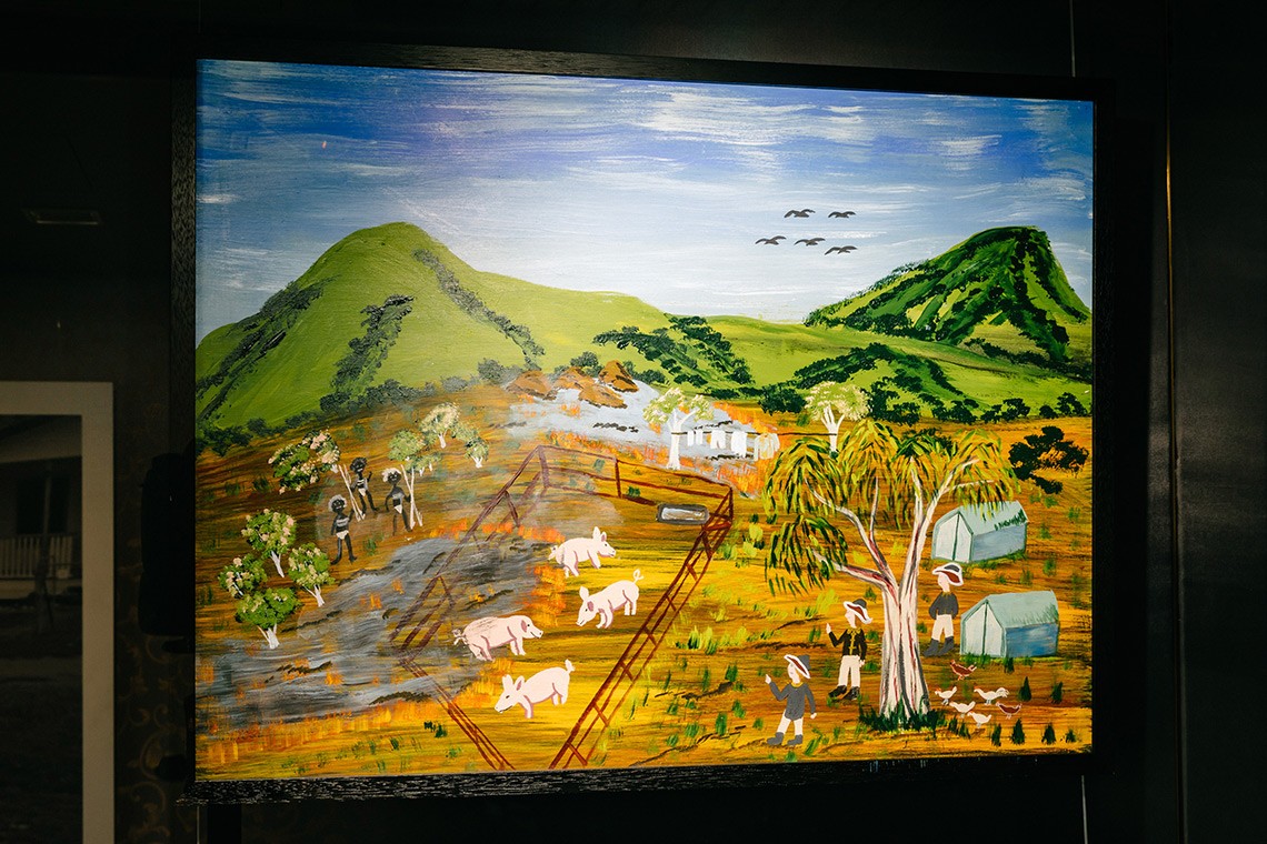 The painting, ""Burning the Pigs"" depicts the First Nations’ oral history of Captain Cook’s landing at Endeavour River.  The painting is acrylic on plywood and depicts the green hills of the endeavour river in the background.  The foreground features a burned pig pen  containing four pigs, with Bama people on the left and Captain Cook's men on the right with their tents and chickens.