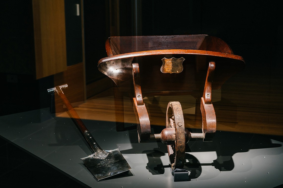 The presentation nickel silver spade and timber wheelbarrow were presented to Arthur Palmer, former Queensland Premier at a ceremony held to mark the turning of the first sod of the Sandgate Railway in 1881.  The spade is engraved in large script on the front and is attached to a timber shaft which has a plain T shaped nickel handle.  The model wheelbarrow is constructed from red cedar. 