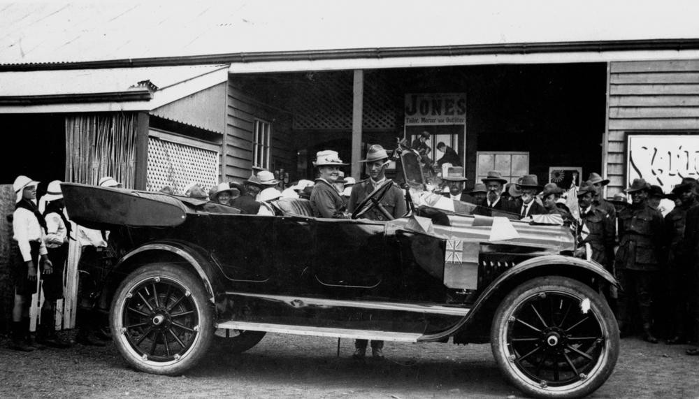 Image of Mrs Wheeler sitting in car in front of buildings with a group of children and adults spread out behind the car ca1919