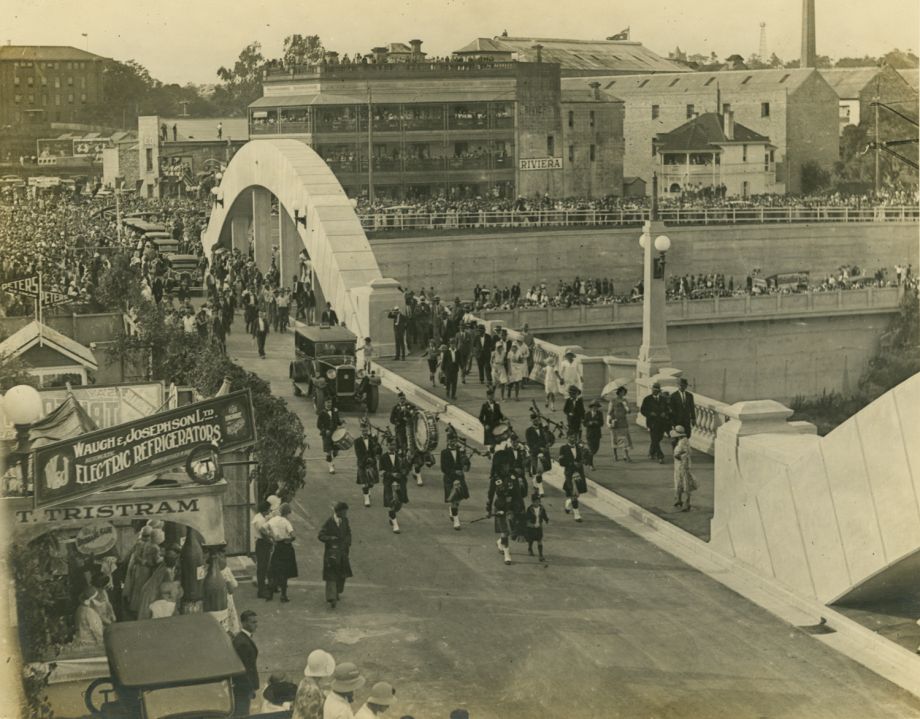 Photo taken as car of the Governor, His Excellency Sir John Goodwin, leads the procession over the Bridge behind the musicians playing bagpipes, after the blue ribbon has been cut. Stalls for the festivities include Tristrams and Peters, seen in left of photo, and other gaily decorated stalls. Riviera Hotel seen in background on north side of river. People crowd the balconies to watch the procession.