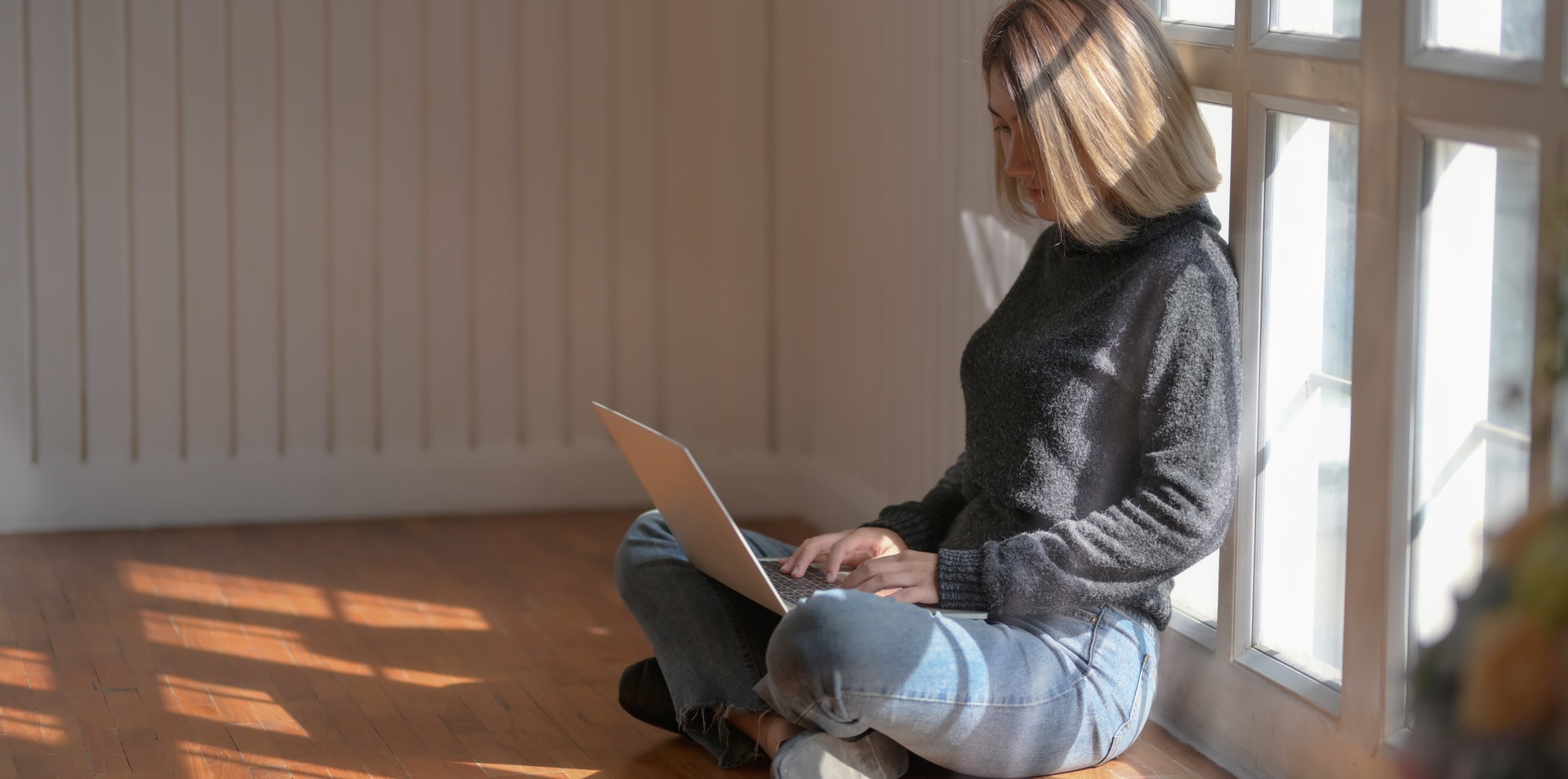 Woman in grey sweater is sitting on the floor and looking at a laptop